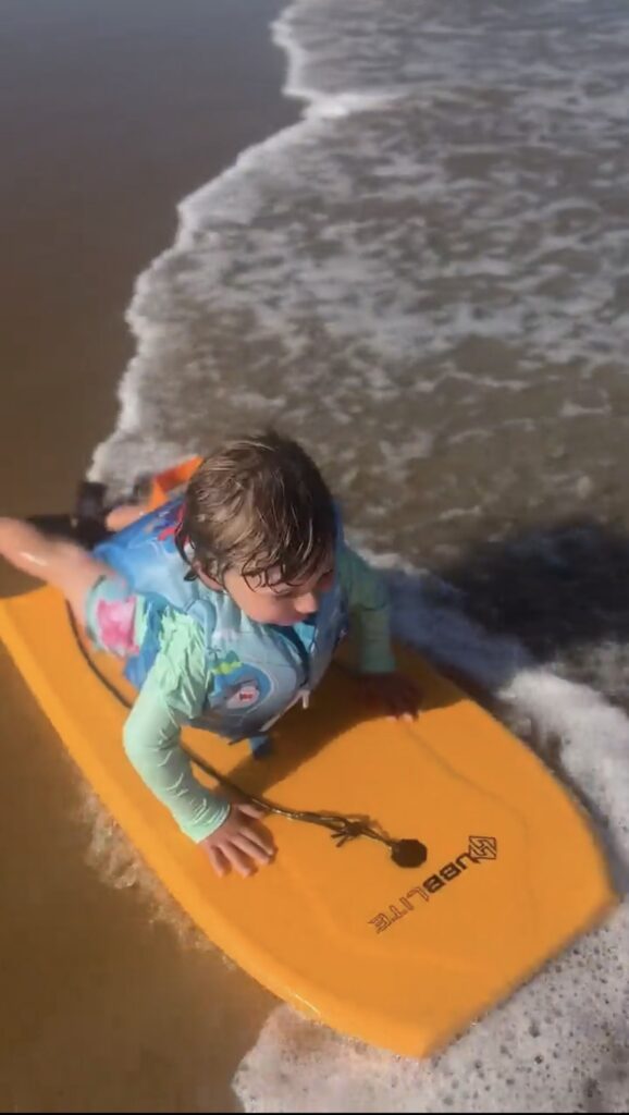 A young child wearing a blue life jacket lies on a yellow boogie board at the edge of the shore, with a small wave washing up on the sand beside them.