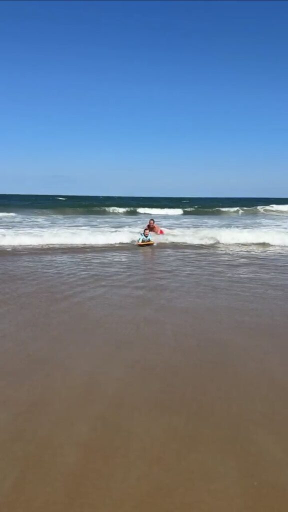A person rides a surfboard on shallow water near the shore, with waves and a bright blue sky in the background. The sandy beach stretches into the foreground.