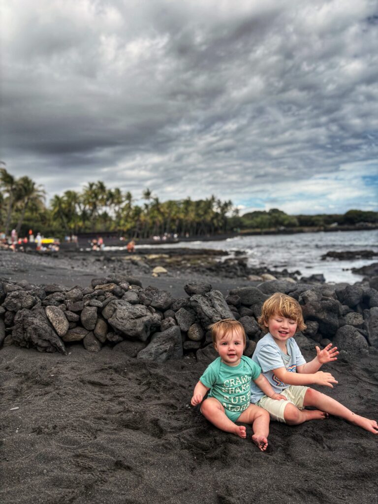 Two young children sit smiling on black sand by a rocky wall at a beach. Palm trees and people are visible in the background under a cloudy sky.