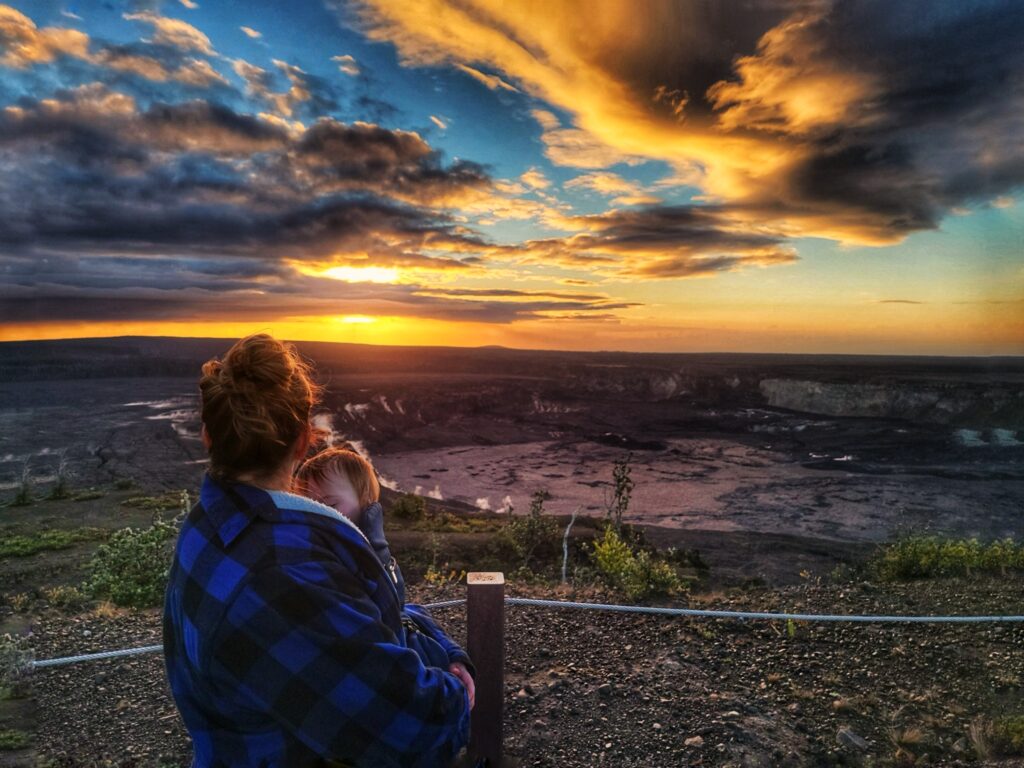 A person in a blue plaid jacket holds a child while watching a dramatic sunset over a rocky, expansive landscape with clouds lit in orange and yellow hues.