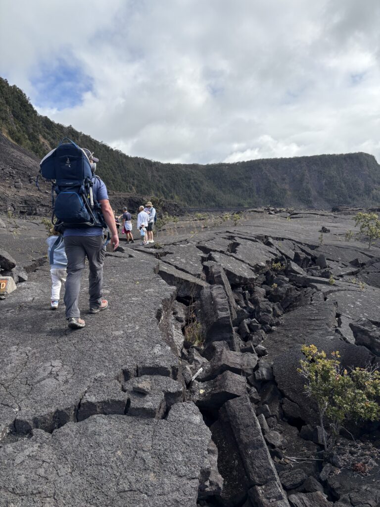 A group of hikers, including a person carrying a child in a backpack carrier, walk along cracked volcanic rock with green hills in the background under a partly cloudy sky.
