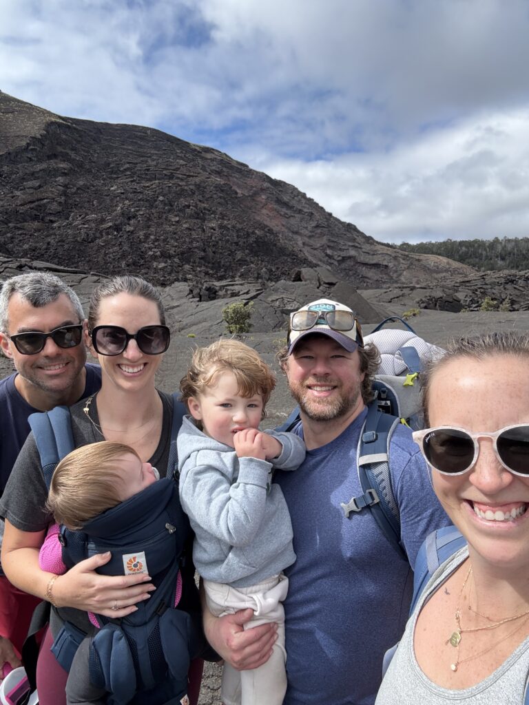 A group of five adults and two small children pose for a selfie while hiking on a rocky, mountainous landscape under a partly cloudy sky. Everyone is smiling, wearing casual outdoor clothes and sunglasses.