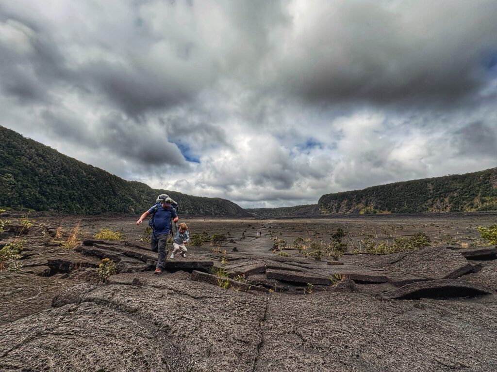 Two people walk across cracked volcanic rock under a cloudy sky, surrounded by green ridges and sparse vegetation in a wide volcanic crater landscape.