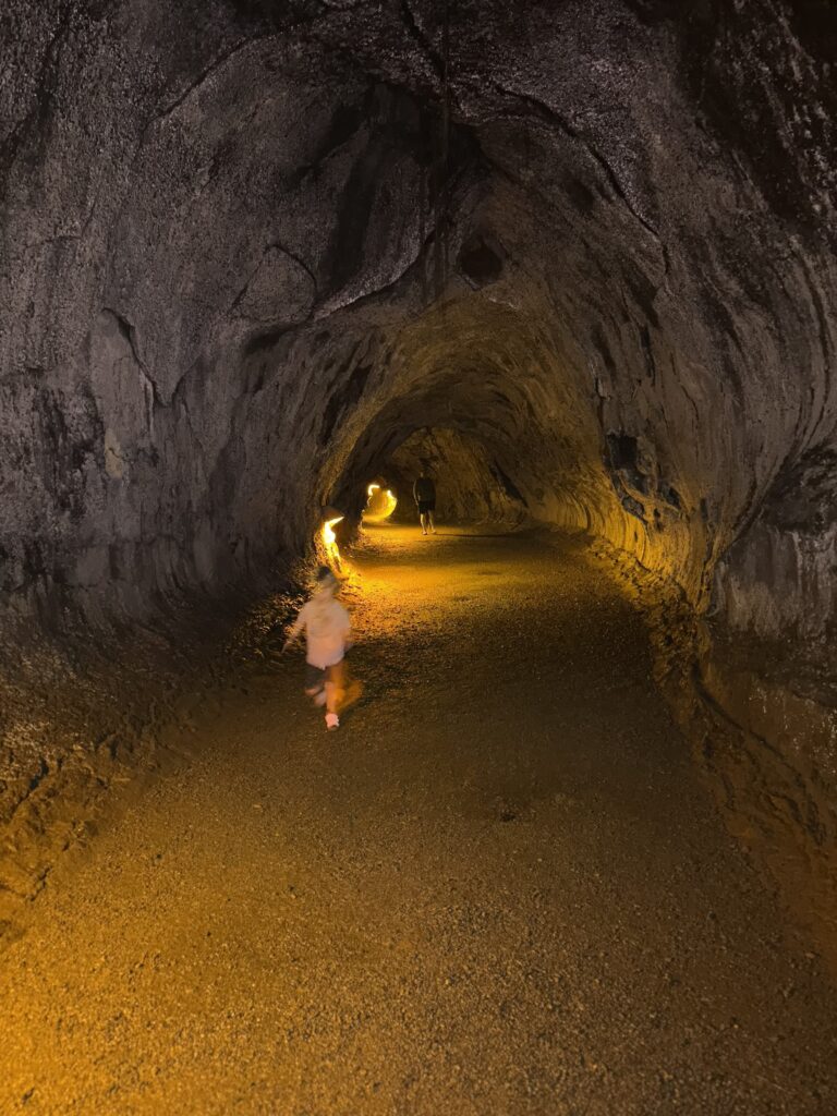 A child runs through a dimly lit lava tube cave with textured rock walls and a curved ceiling, while another person stands in the distance under warm yellow lights.