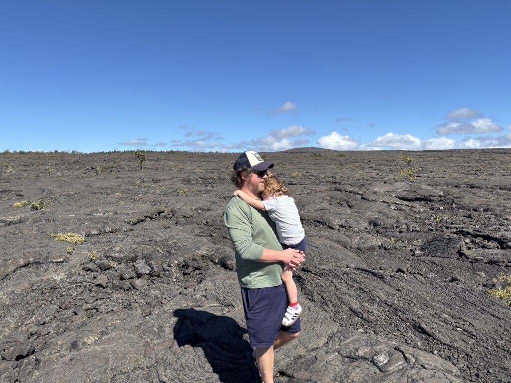 A man wearing a cap holds a small child in his arms while standing on a vast, rocky lava field under a clear blue sky with scattered clouds. Sparse vegetation is visible in the background.