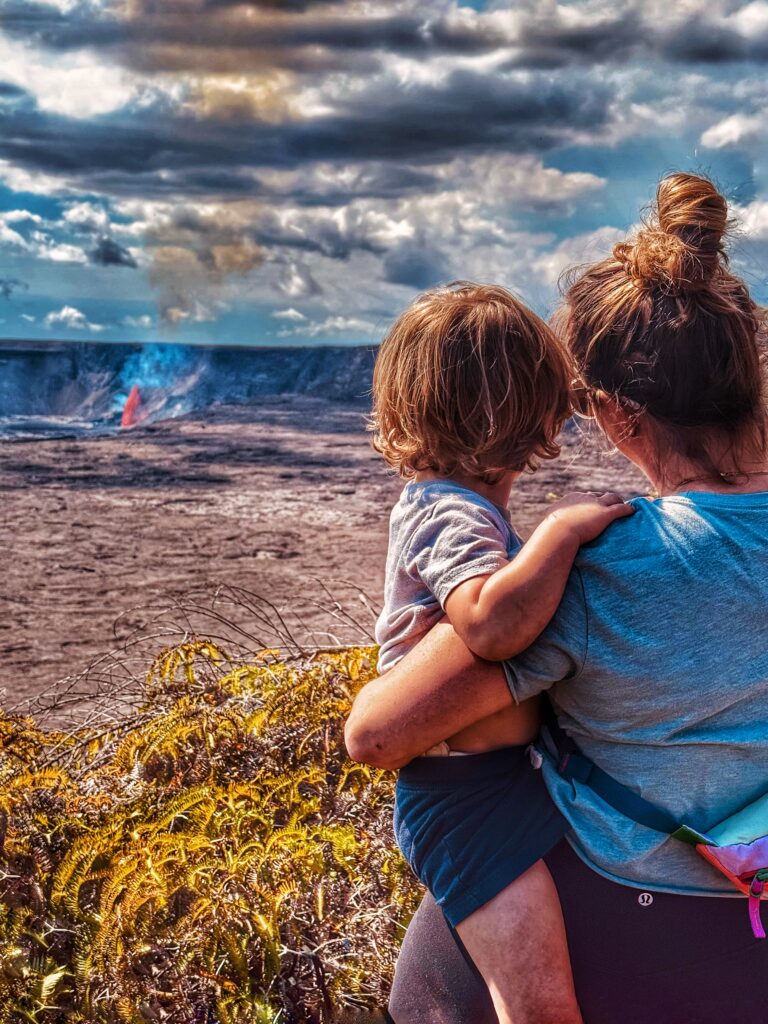 A woman holding a small child looks out over a volcanic landscape with smoke rising and a hint of red lava in the distance under a dramatic, cloudy sky. Yellowish-green plants are in the foreground.
