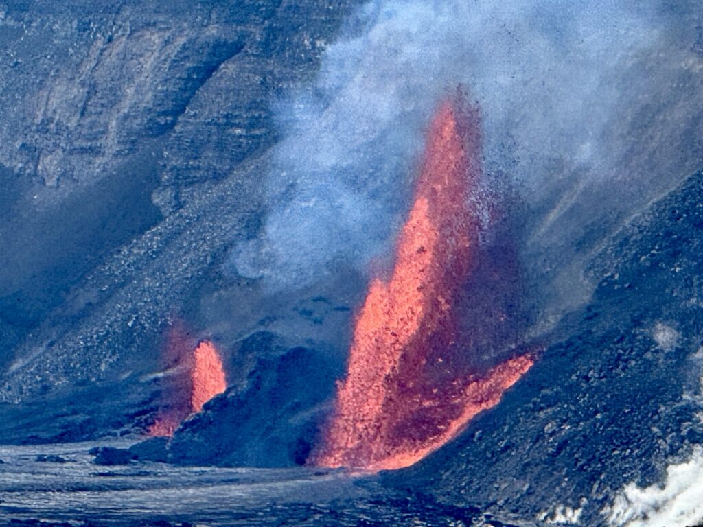 Bright orange lava fountains erupt from a rocky volcano slope, sending smoke and ash into the air. Dark volcanic rock surrounds the flow, contrasting with the vivid color of the erupting lava.