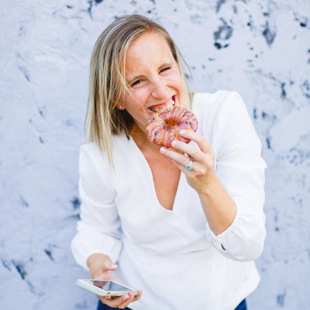 A woman with blonde hair, wearing a white blouse, smiles while holding a pink glazed donut close to her mouth. She holds a smartphone in her other hand and stands in front of a light purple textured wall.