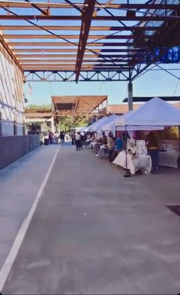 Outdoor market scene with white vendor tents lined up on the right side of a wide covered walkway, people browsing booths, and blue sky visible above through open steel beams.
