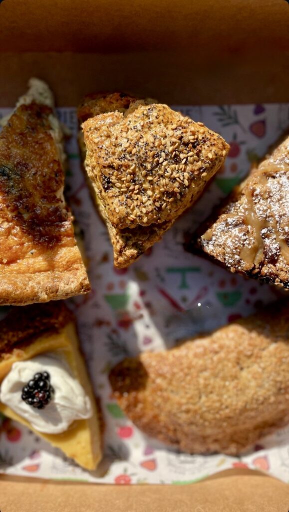 A close-up of five assorted baked goods, including scones, a tart with whipped cream and a blackberry, and other pastries, all arranged on a colorful, patterned tray. Sunlight highlights their textures and toppings.