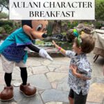 A young boy high-fives Mickey Mouse, dressed in a Hawaiian shirt, at an outdoor breakfast with tropical plants and rocks. Text overlay reads “Aulani Character Breakfast,” highlighting this magical morning experience.