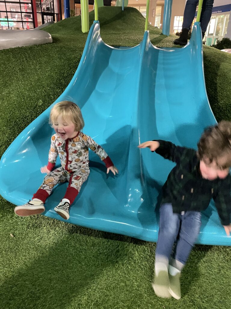 Two young children are playing on a bright blue double slide with artificial grass around it. One child is wearing patterned pajamas and sneakers, while the other is in jeans and a dark jacket, both sliding down and smiling.
