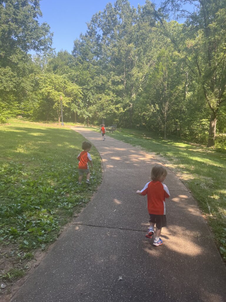 Three young children wearing red shirts walk and play on a winding paved path in a green, sunlit park surrounded by trees and grass.