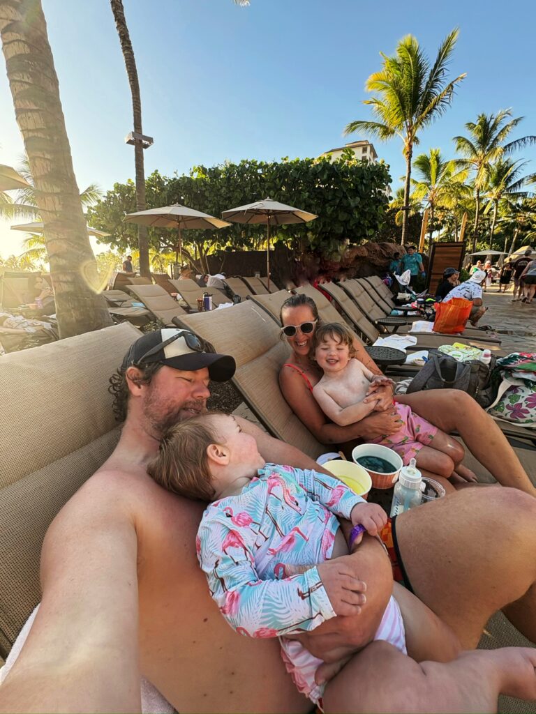 A family of four relaxes on poolside lounge chairs in swimsuits, surrounded by palm trees and sun umbrellas, with blue skies overhead. Two children sit with their parents, smiling and enjoying drinks.