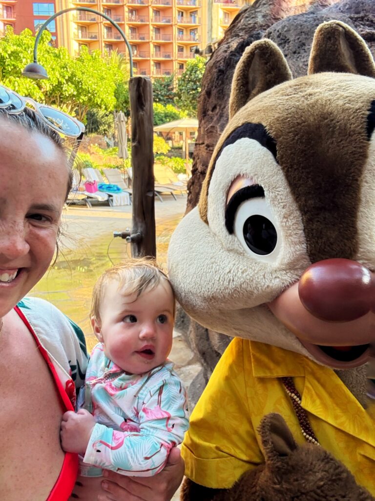 A smiling woman holds a baby who looks up at Chip, a large costumed chipmunk character in a yellow shirt, at an outdoor resort with greenery and buildings in the background.