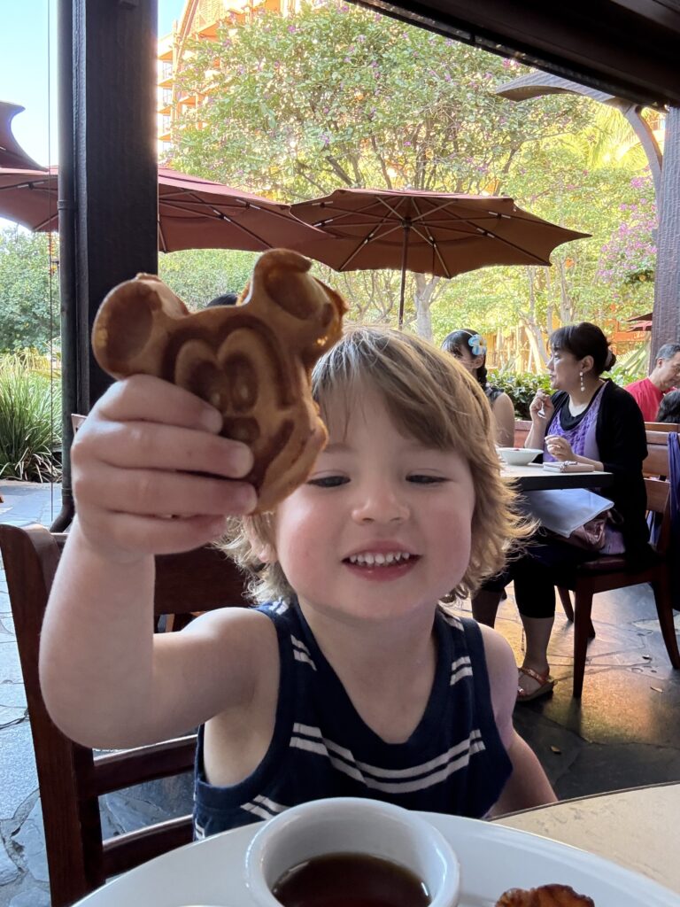 A smiling young child holds up a Mickey Mouse-shaped waffle at an outdoor restaurant, with people dining and trees with pink flowers in the background.