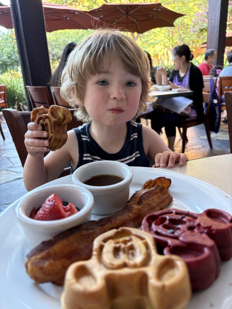 A young child sits at an outdoor table, holding a Mickey Mouse-shaped waffle. In front of them is a plate with more Mickey waffles, a strip of bacon, syrup, and strawberries. Other diners are visible in the background.