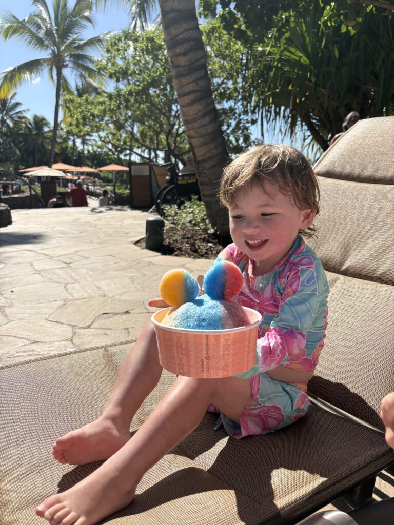 A young child in a colorful swimsuit sits on a lounge chair outdoors, smiling and holding a bowl of rainbow shaved ice. Palm trees and sunny weather suggest a tropical location.