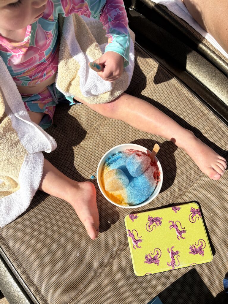 A child in a colorful swimsuit sits on a lounge chair with a towel, enjoying a bowl of rainbow shaved ice and a yellow tray with purple seahorse designs. Sunlight casts shadows on the scene.