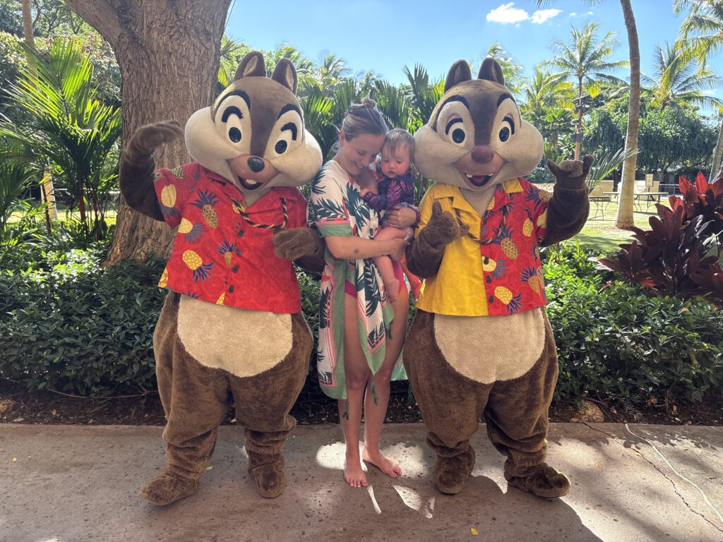 A woman holding a baby stands between two people in Chip and Dale costumes wearing Hawaiian shirts, surrounded by lush greenery and palm trees on a sunny day.