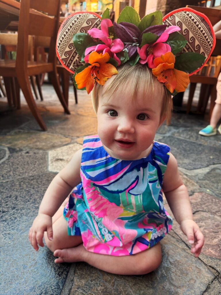 A smiling baby sits on a stone floor wearing a colorful, tropical-patterned dress and a floral headband with large decorated mouse ears. Wooden chairs and another person’s legs are visible in the background.