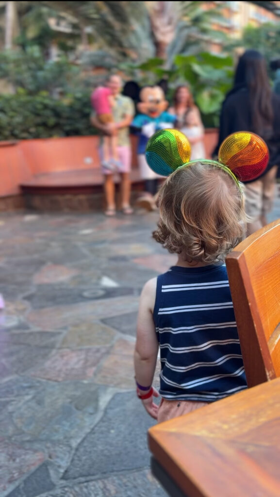 A small child with curly hair and rainbow Mickey Mouse ears sits on a bench, looking toward people posing with a costumed character in the background. The scene is outdoors with stone pavement and greenery.