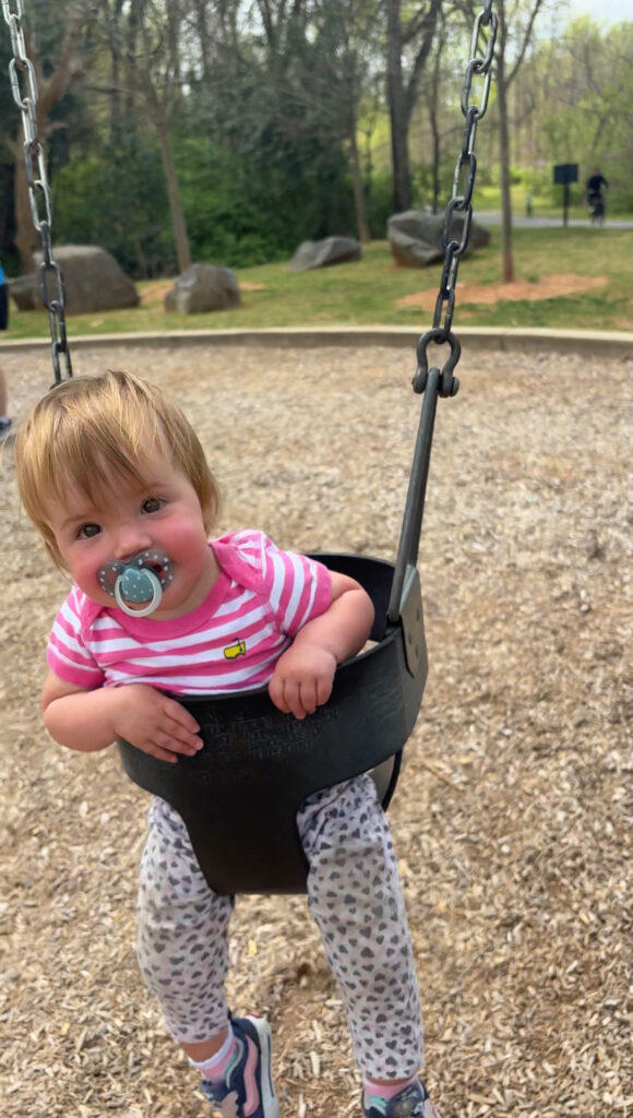 A smiling baby with a pacifier, wearing a pink striped shirt and patterned pants, sits in a bucket swing at a playground. Trees, rocks, and wood chips are visible in the background.