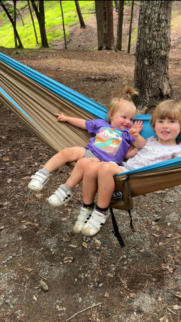 Two young children sit together in a brown and blue hammock outdoors in a wooded area. One child wears a purple shirt and white sandals, and the other wears a white shirt, both smiling and relaxing among trees.