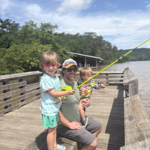 A man kneels on a wooden dock by a lake, smiling with two young children. One child holds a yellow fishing pole while the other sits on the mans knee. Trees and blue sky are in the background.