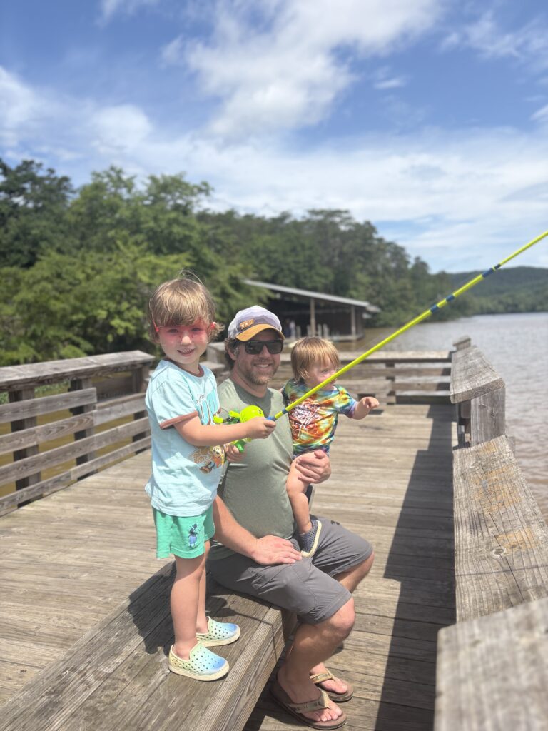 A man kneels on a wooden dock by a lake, smiling with two young children. One child holds a yellow fishing pole while the other sits on the mans knee. Trees and blue sky are in the background.