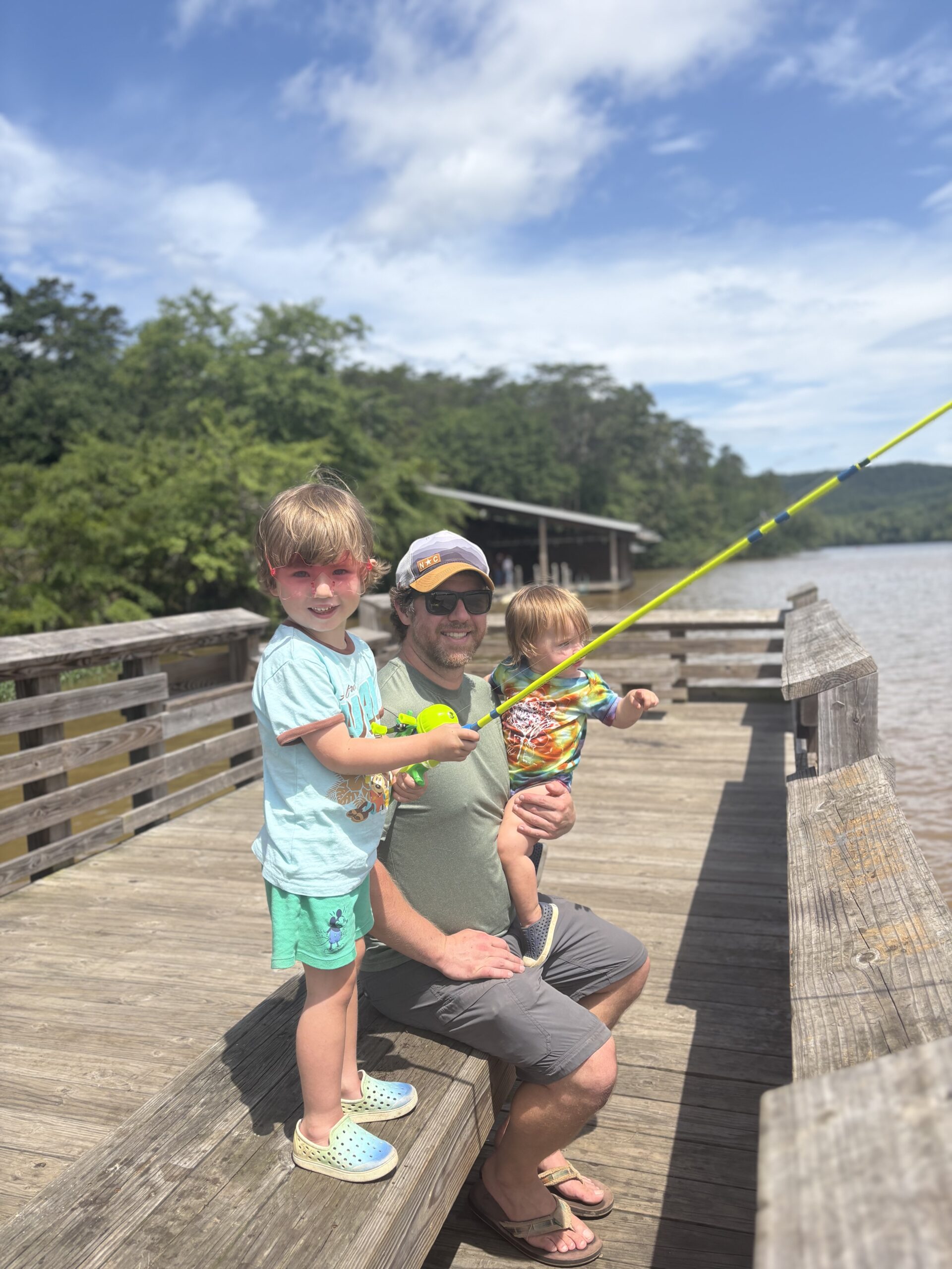 A man kneels on a wooden dock by a lake, smiling with two young children. One child holds a yellow fishing pole while the other sits on the mans knee. Trees and blue sky are in the background.