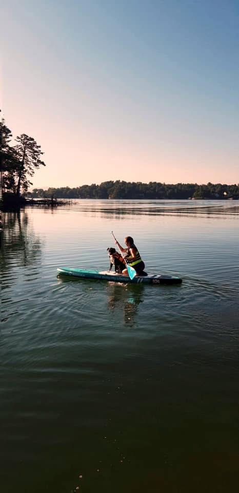 A person wearing a life vest paddles a kayak on a calm lake with a dog seated at the front. Trees and a clear sky are visible in the background.