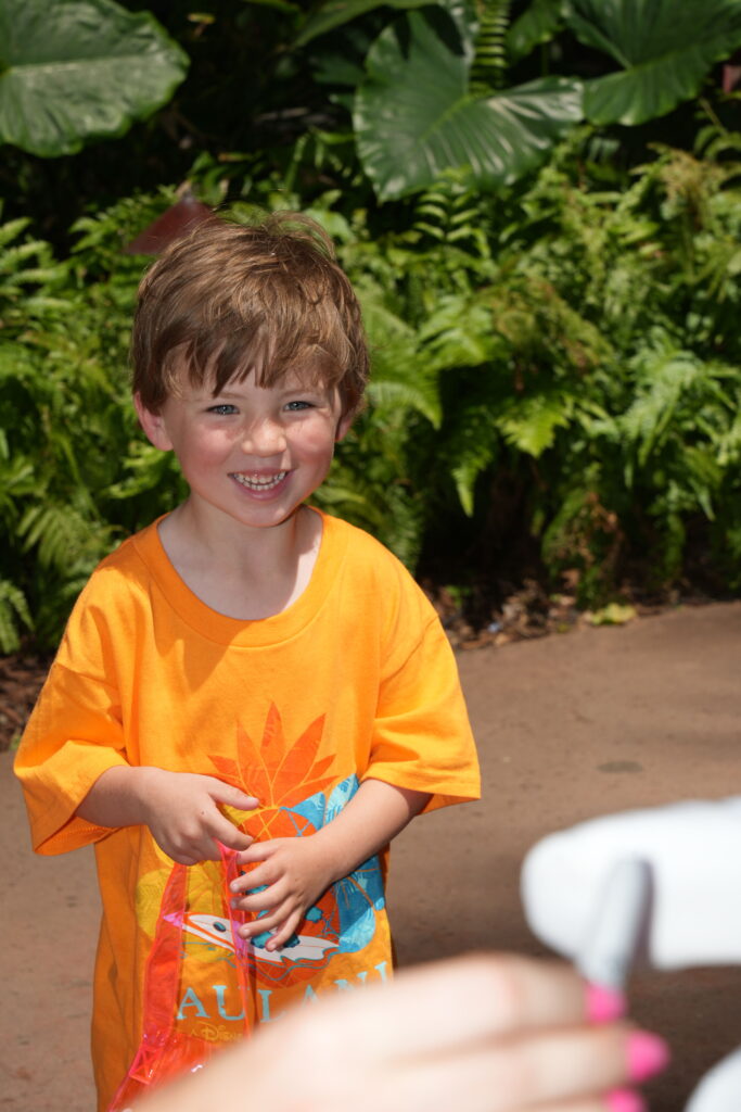 A young boy with brown hair, wearing an orange T-shirt, smiles outdoors while holding a red bag. Green plants and ferns are in the background; a blurred hand appears in the foreground.