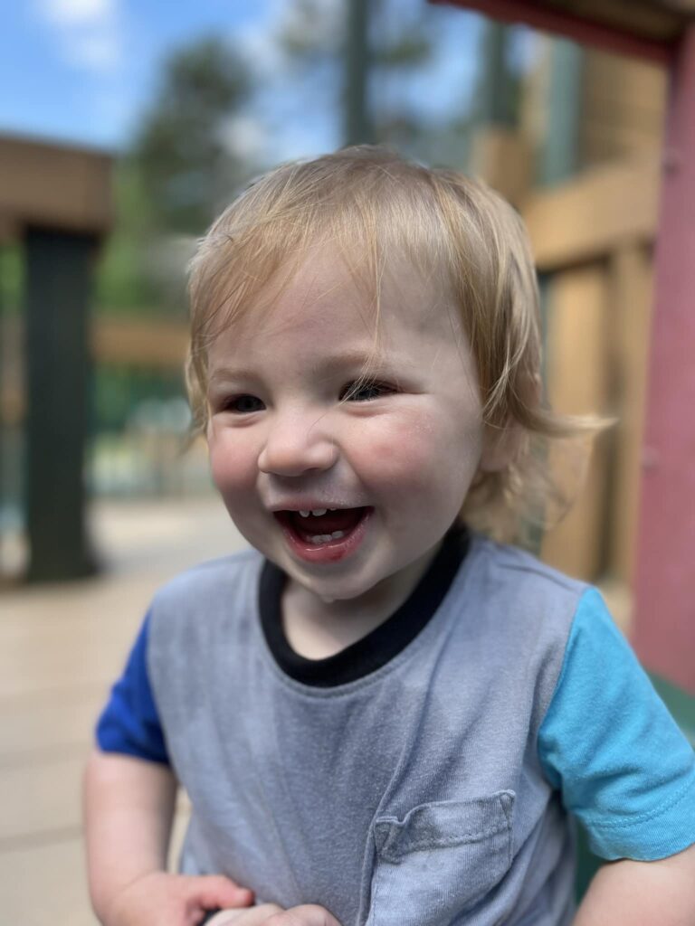 A young child with blonde hair and a big smile stands outdoors at Charlotte Parks, wearing a gray and blue shirt. The background is softly blurred, showing greenery and wooden structures.