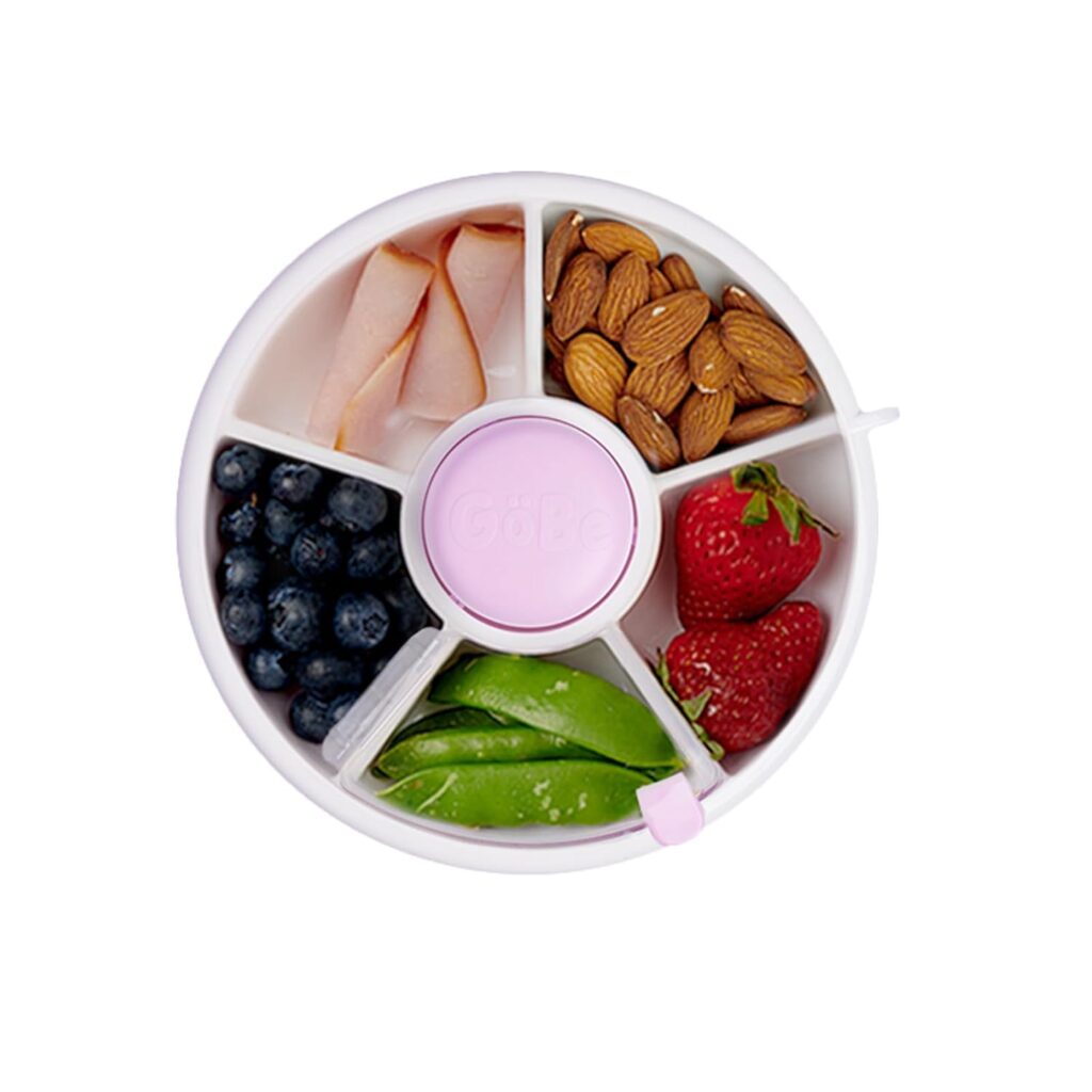 A round, sectioned snack container holds sliced turkey, almonds, strawberries, blueberries, and snap peas, with a central pink lid.