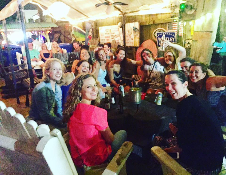 A group of people sit around a table at a lively indoor-outdoor venue, smiling and posing for the camera. The setting, inspired by the vibrant spirit of North Carolina beaches, features colorful decor and string lights for a festive atmosphere.