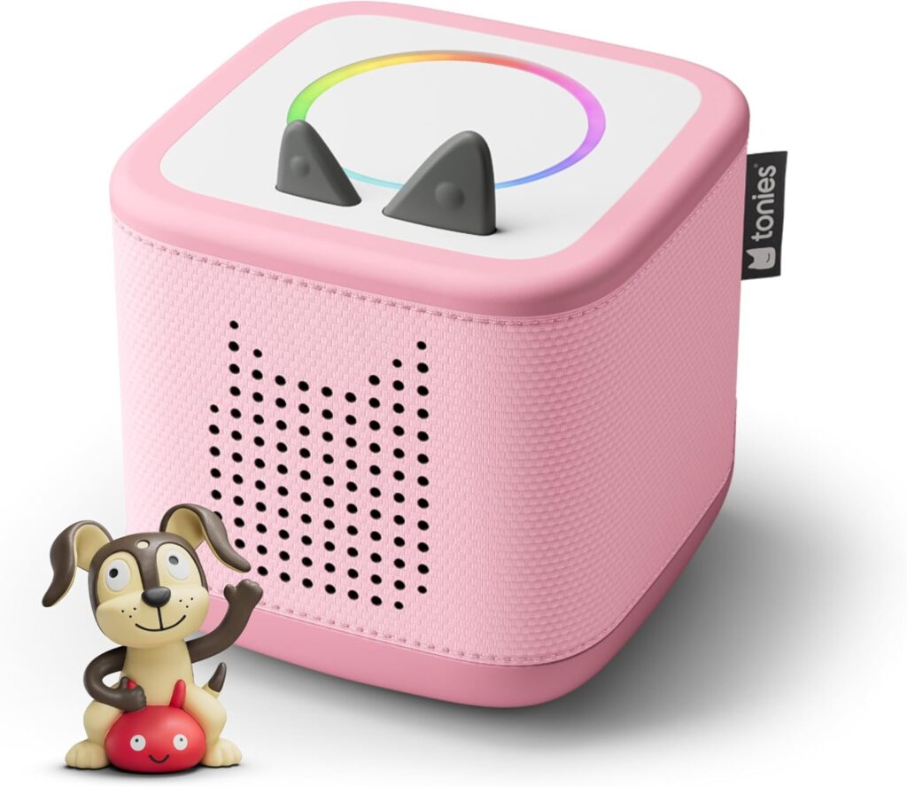 A pink Toniebox audio player with cat ear-shaped controls on top and a speaker in the shape of a cats face. In front is a small dog figurine holding a red object.