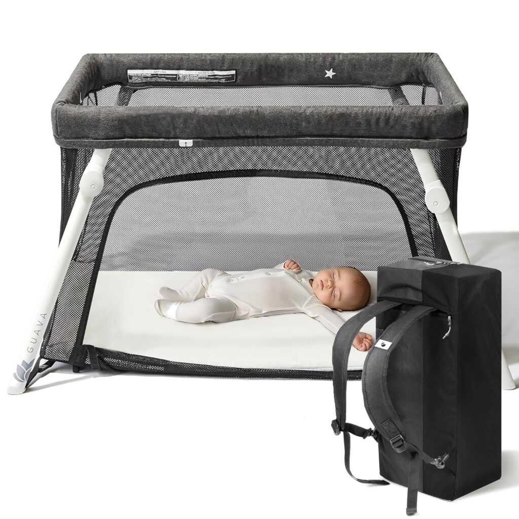 A baby sleeps in a gray mesh portable crib with white legs. A black carrying case with backpack straps is placed in front of the crib.