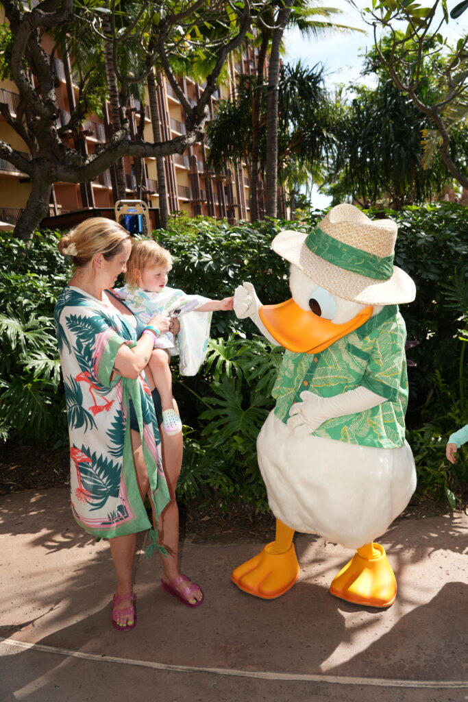 A woman holding a young child greets a person in a Donald Duck costume wearing a green shirt and straw hat, outdoors near lush greenery and palm trees.