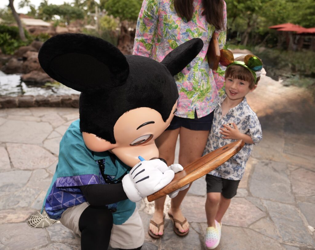 A person in a Mickey Mouse costume signs an autograph for a smiling young child in vacation attire, while an adult stands nearby; they are outdoors on a stone path with greenery in the background.