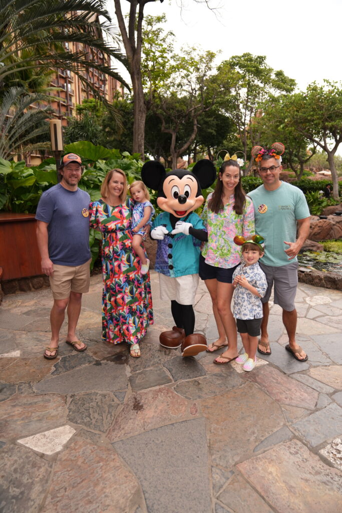 A group of five people, including two young children, stands outdoors with Mickey Mouse. They are smiling and dressed in colorful vacation attire, surrounded by lush greenery and tropical plants.