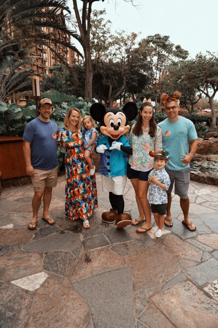 A family of six poses outdoors on a stone pathway with Mickey Mouse, who is dressed in a blue Hawaiian shirt. Lush greenery and trees are in the background, creating a tropical atmosphere.
