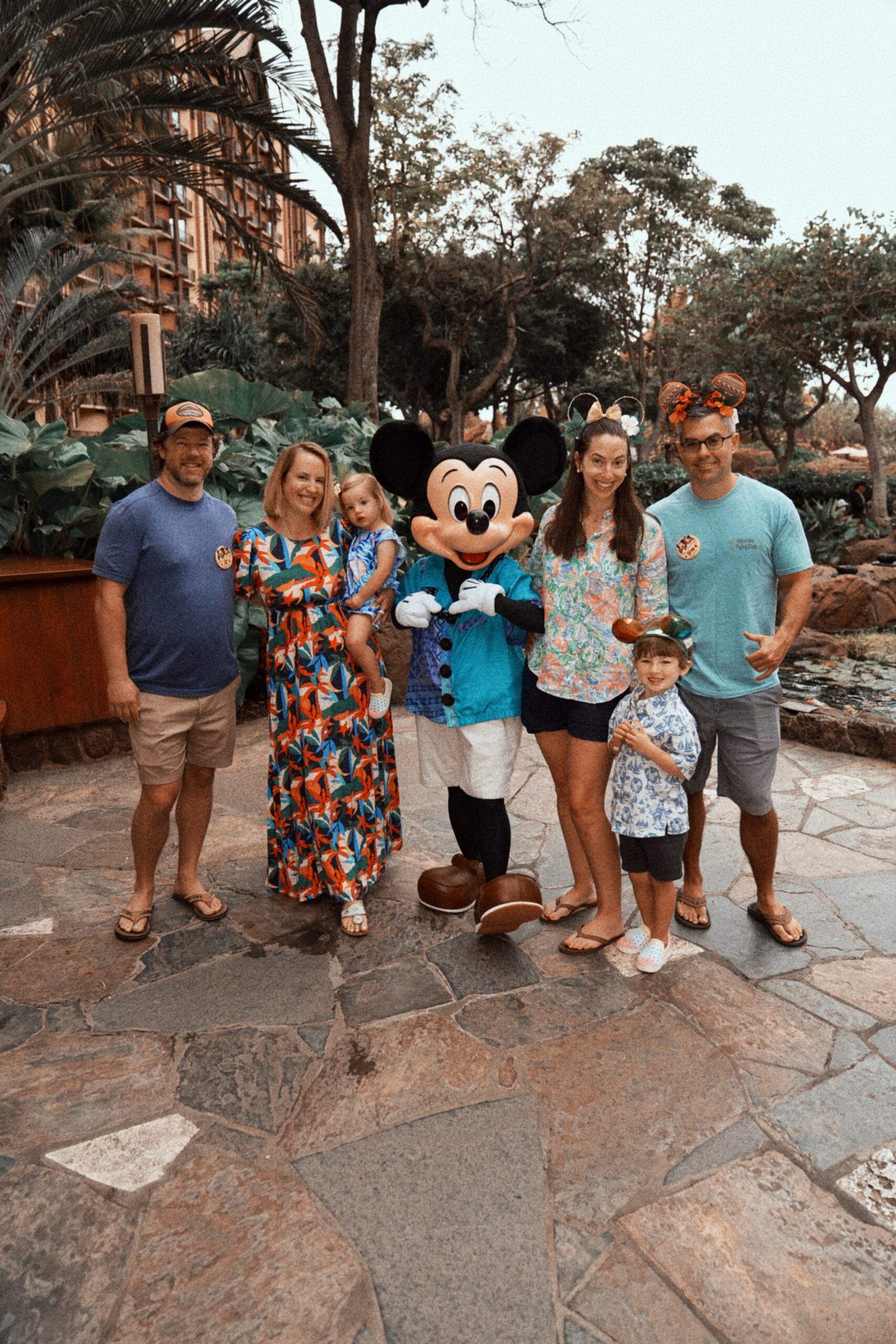 A family of six poses outdoors on a stone pathway with Mickey Mouse, who is dressed in a blue Hawaiian shirt. Lush greenery and trees are in the background, creating a tropical atmosphere.