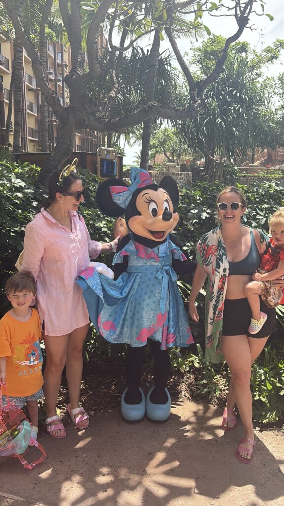 Two women and two children pose outdoors with someone in a Minnie Mouse costume wearing a blue and pink dress. The group is smiling in front of green foliage under bright sunlight.