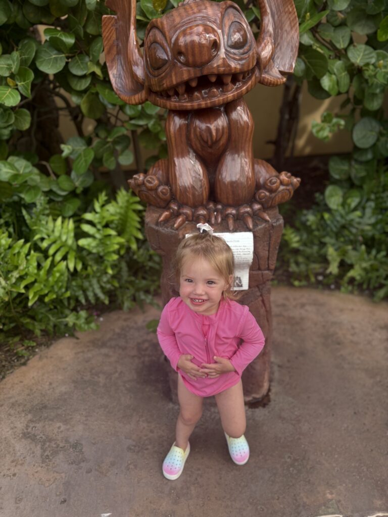 A smiling toddler in a pink outfit stands in front of a carved wooden statue with large ears and a wide mouth, surrounded by lush green plants.