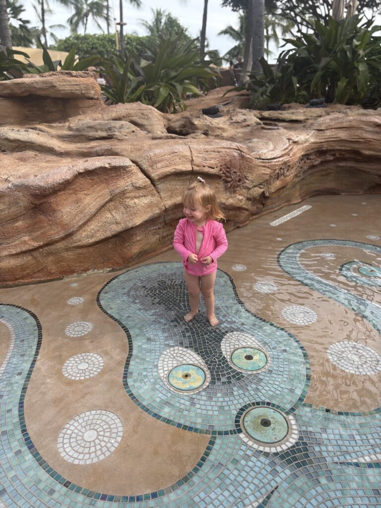 A young child wearing a pink swimsuit stands barefoot in shallow water on a decorative tiled surface, with large faux rock formations and tropical plants in the background.