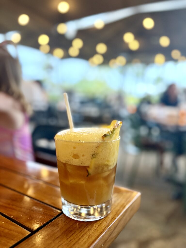 A frothy tropical cocktail garnished with a pineapple slice sits on a wooden table at an outdoor restaurant, with blurred people and string lights in the background.