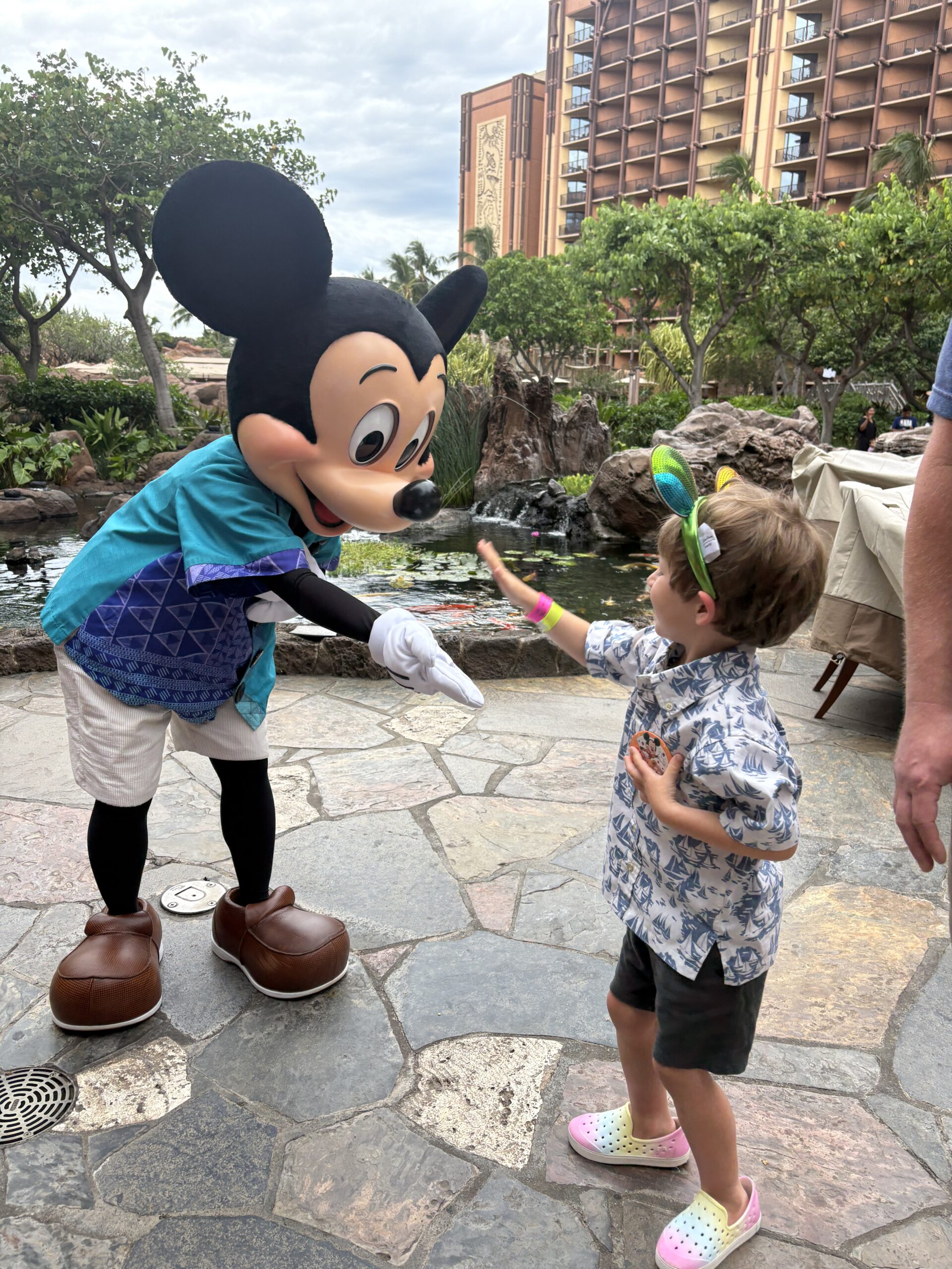 A child wearing a Hawaiian shirt and colorful shoes high-fives a person in a Mickey Mouse costume dressed in island attire. They are outdoors on a stone path with lush greenery and a hotel in the background.