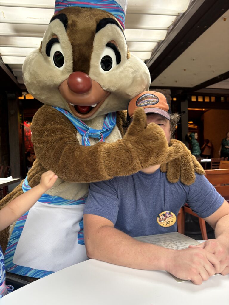 A costumed chipmunk character hugs a seated man wearing a baseball cap and blue shirt at a restaurant. The man holds a menu, and a childs arm is visible on the left. The chipmunk gives a thumbs-up.