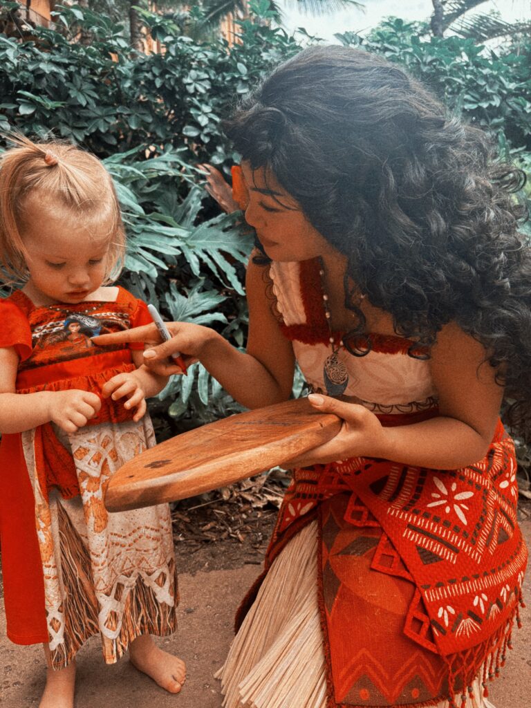 A woman in a red and white Polynesian-inspired outfit kneels and smiles as she interacts with a young girl in a matching dress, both surrounded by lush greenery. The woman holds a wooden board and points at the girls hand.